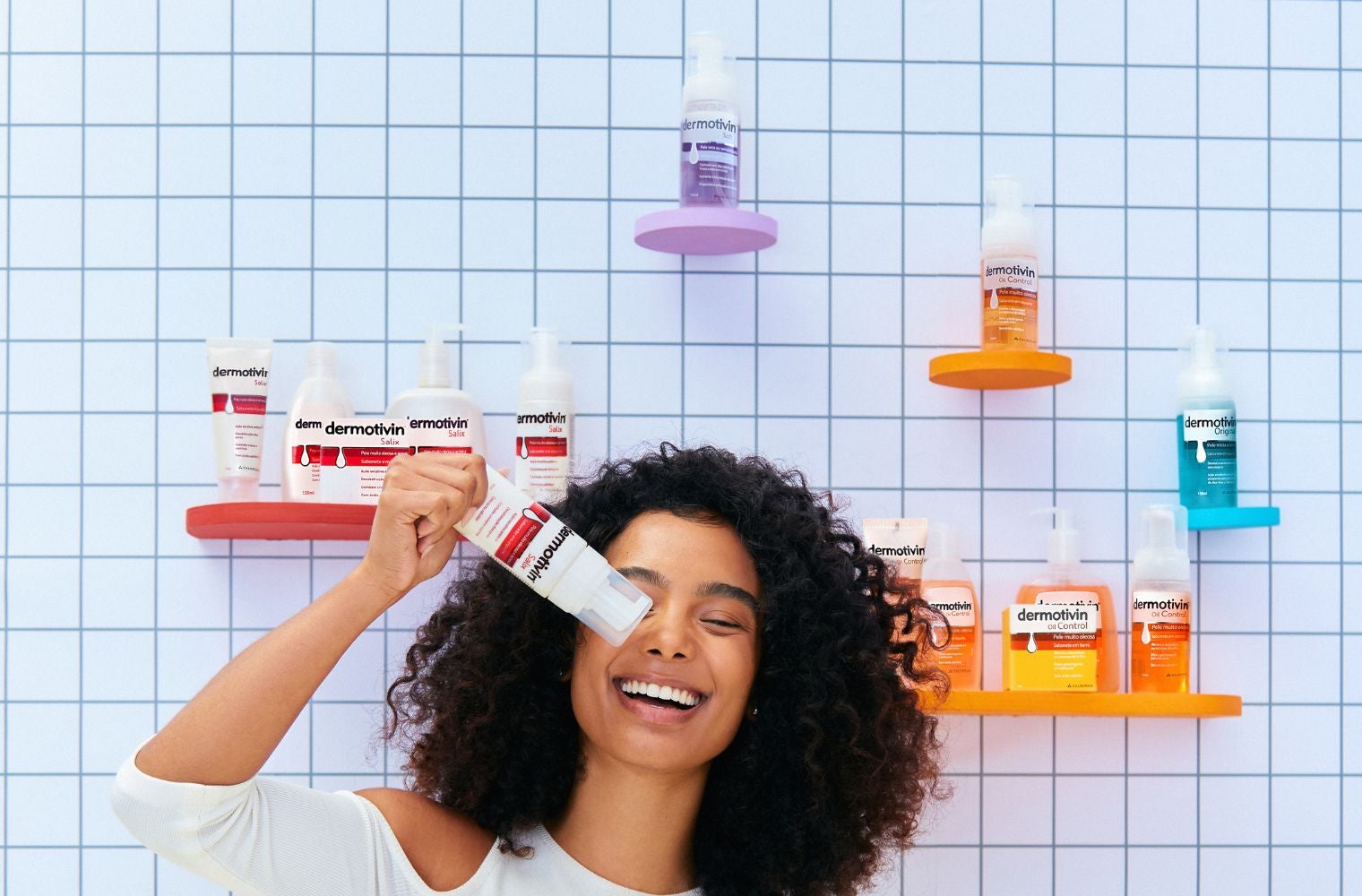 Mulher negra sorrindo e segurando sabonete em espuma Dermotivin Salix. Ao fundo, prateleiras com as linhas de Dermotivin Oil Control e Salix e as espumas de Dermotivin Soft e Original.