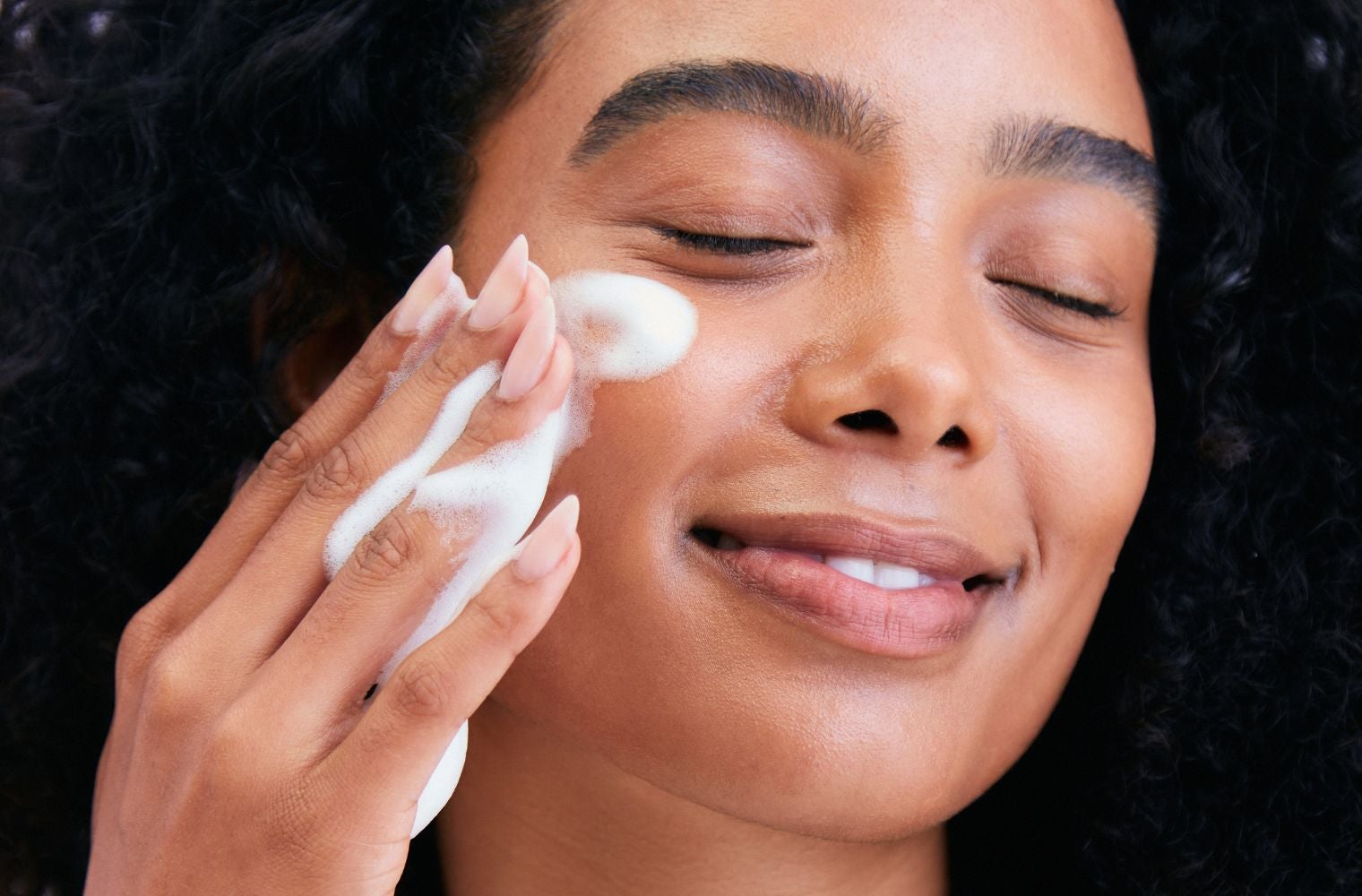 Mulher negra sorrindo e aplicando, no rosto, um sabonete em espuma.