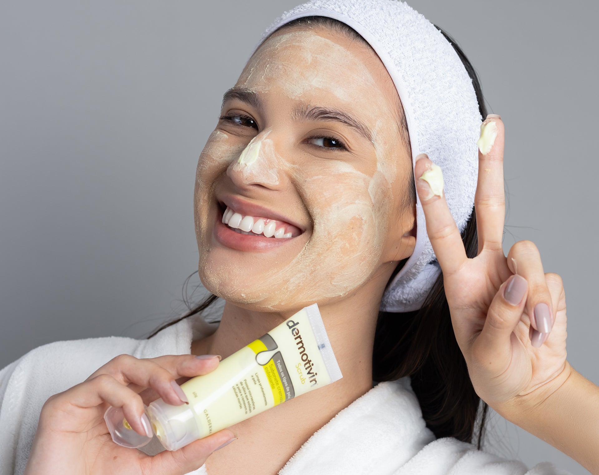 Uma mulher sorridente, vestindo um roupão branco e uma faixa de cabelo, aplica o Dermotivin Scrub no rosto. Ela segura o tubo do produto em uma mão e faz o sinal de "paz e amor" com a outra, transmitindo uma sensação de bem-estar e cuidado com a pele. A imagem destaca a aplicação do produto de esfoliação facial, mostrando a textura cremosa e a cobertura uniforme na pele. A expressão feliz da modelo reforça a ideia de que a esfoliação facial com Dermotivin Scrub é um momento agradável e parte de uma rotina d