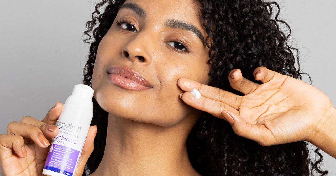 Mulher negra segurando Hidratante Microbioma Equilibrado Dermotivin Benzac em uma das mãos. O produto está em destaque, com sua embalagem branca e detalhes em lilás. 