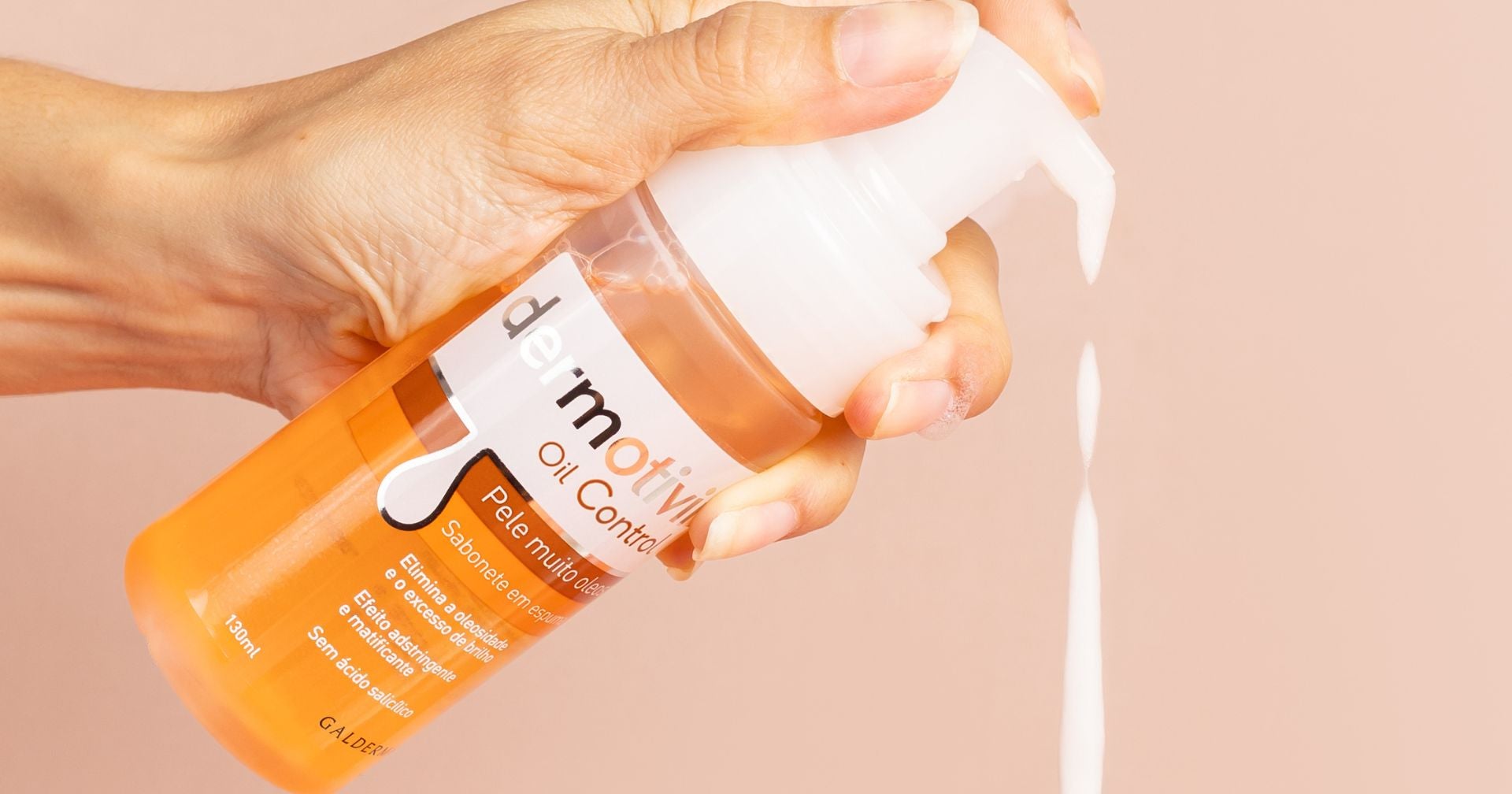 Uma mão segurando um frasco de produto para a pele da marca Dermotivin Oil Control, com espuma branca saindo do bico dosador. O fundo é amarelo, destacando o frasco e a textura do produto.