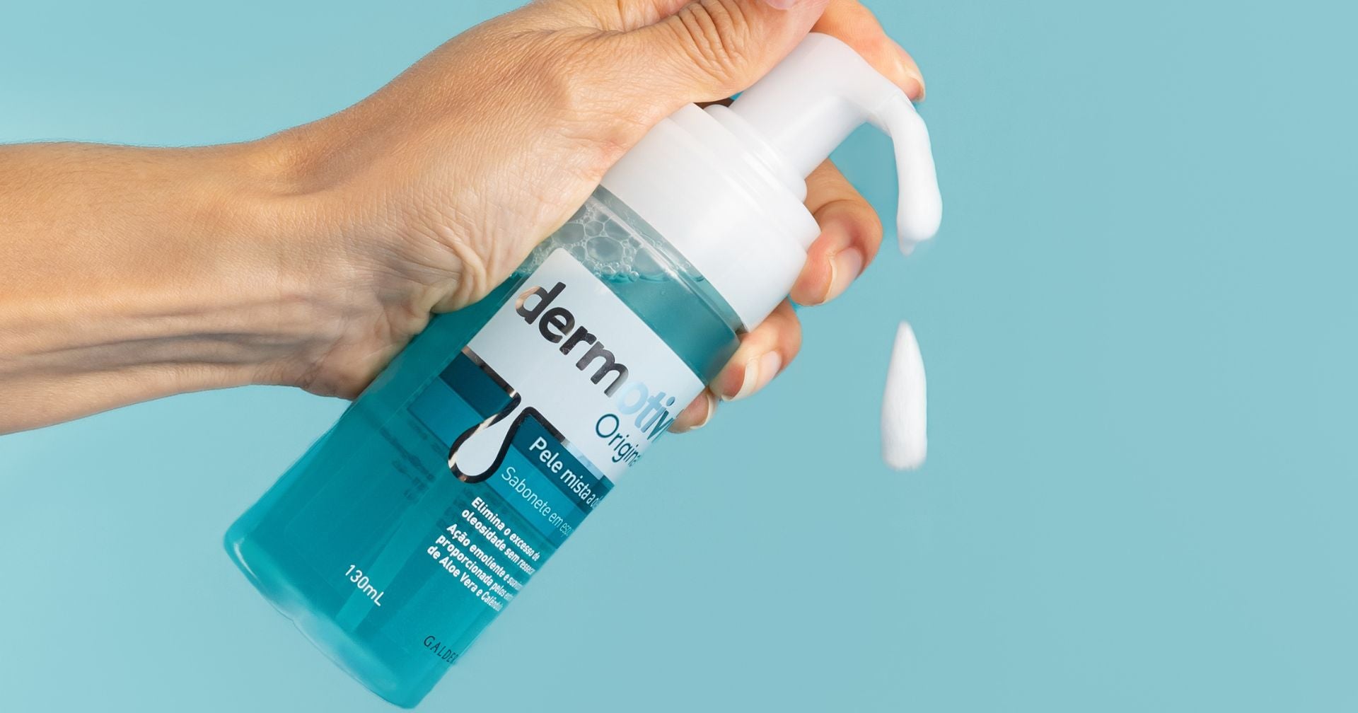Uma mão segurando um frasco de produto para a pele da marca Dermotivin Original, com espuma branca saindo do bico dosador. O fundo é verde, destacando o frasco e a textura do produto.
