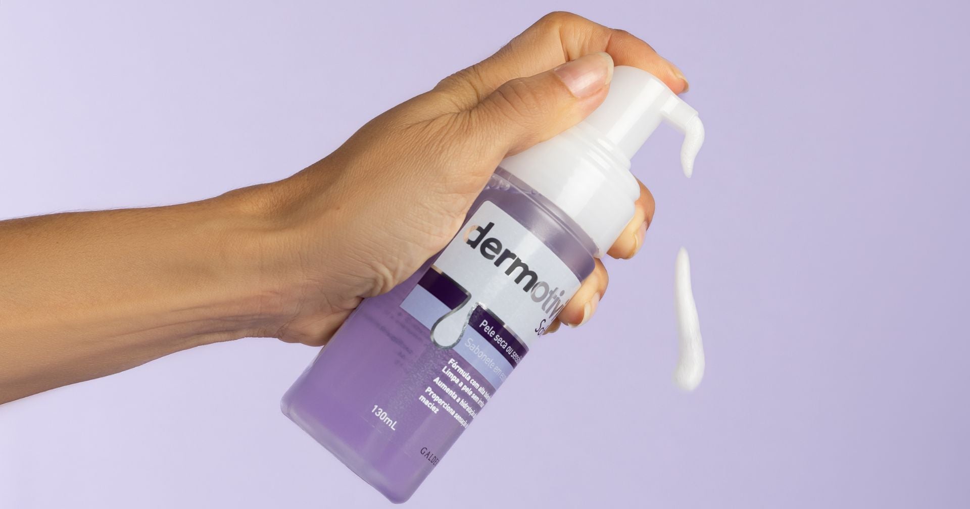 Uma mão segurando um frasco de produto para a pele da marca Dermotivin Soft, com espuma branca saindo do bico dosador. O fundo é lilás, destacando o frasco e a textura do produto