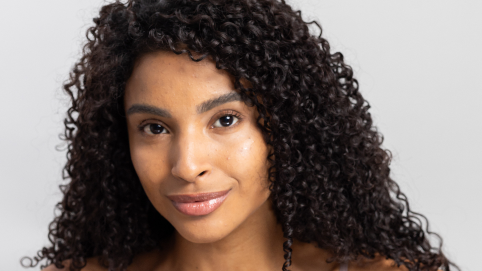 Mulher jovem com cabelos cacheados escuros e pele radiante sorrindo suavemente, demonstrando uma pele saudável e bem cuidada em fundo neutro.