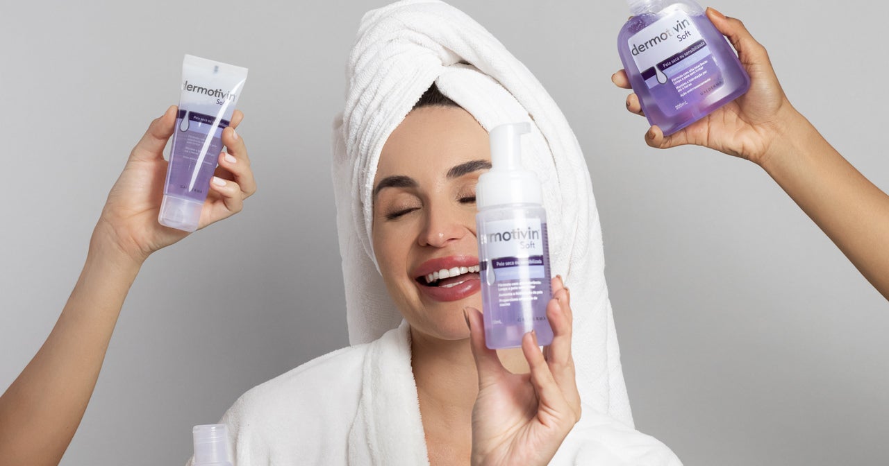Uma mulher com uma toalha enrolada na cabeça está sorrindo e segurando um frasco de produto para cuidados com a pele. Ela está vestindo um roupão branco. Três mãos seguram outros produtos para cuidados com a pele ao redor dela. Os produtos estão todos em embalagens roxas e brancas. O fundo é cinza liso. A imagem geral tem uma estética limpa e brilhante, sugerindo um foco em cuidados com a pele e beleza.