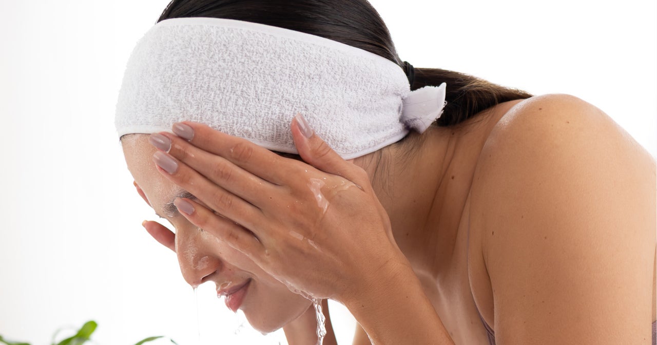Mulher realizando sua rotina de cuidados faciais, lavando o rosto com água sobre uma pia. Ela usa uma faixa branca para prender o cabelo, mantendo-o afastado do rosto durante a limpeza. A imagem mostra o momento da enxágue, com a água escorrendo suavemente pelas mãos posicionadas sobre o rosto, demonstrando a importância da limpeza adequada na rotina de skincare para pele oleosa.