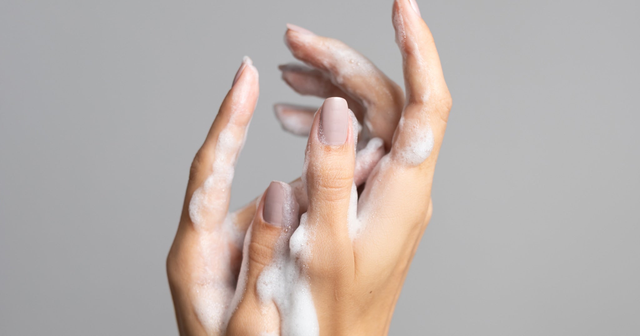 Uma imagem de close-up de duas mãos femininas com unhas pintadas de um tom neutro, lavando-se com sabão, criando uma espuma branca e espessa. As mãos estão posicionadas uma sobre a outra, com os dedos ligeiramente curvados e a espuma cobrindo a maior parte da superfície da pele. O fundo é um cinza sólido e suave, que não distrai das mãos em primeiro plano.