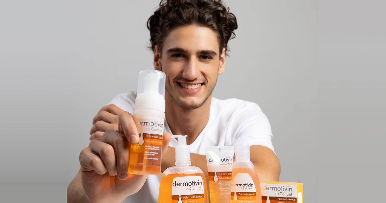 Um jovem com cabelo castanho encaracolado e uma camisa branca sorri para a câmera enquanto segura um frasco de Dermotivin Oil Control, um limpador facial espumoso de cor âmbar. Na frente dele, sobre uma superfície clara, estão dispostos outros produtos da mesma linha Dermotivin Oil Control, incluindo um sabonete líquido em frasco com pump, um tubo, outro frasco de sabonete líquido e uma caixa de sabonete em barra, todos com embalagens nas cores laranja e branco. O fundo é liso e de cor clara.
