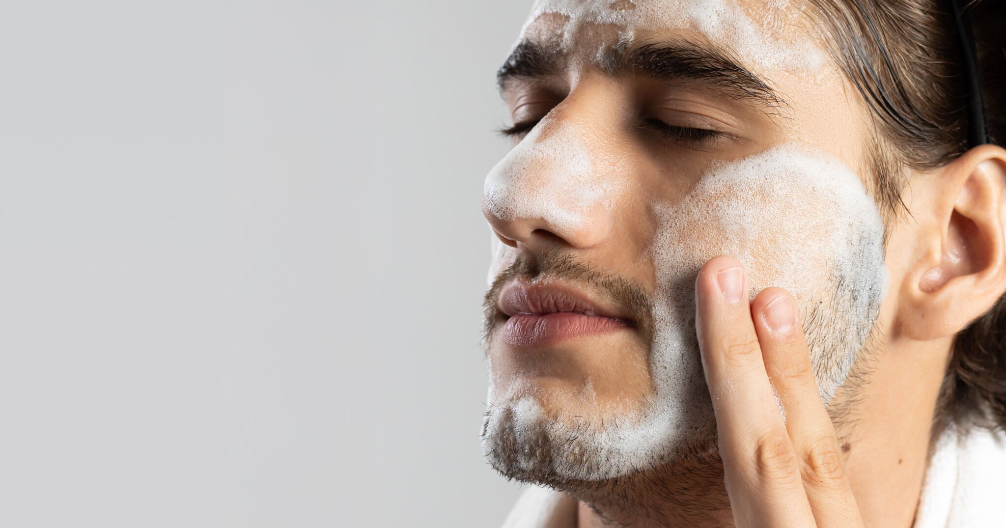 Um close-up de um homem jovem com cabelo escuro e barba por fazer, de perfil, com os olhos fechados, aplicando Espuma de Limpeza no rosto. Ele está usando uma faixa de cabelo preta e uma toalha branca. A espuma branca cobre a testa, bochecha e queixo, enquanto ele usa o dedo indicador e médio da mão direita para espalhar o produto na bochecha. O fundo é um cinza claro e desfocado.