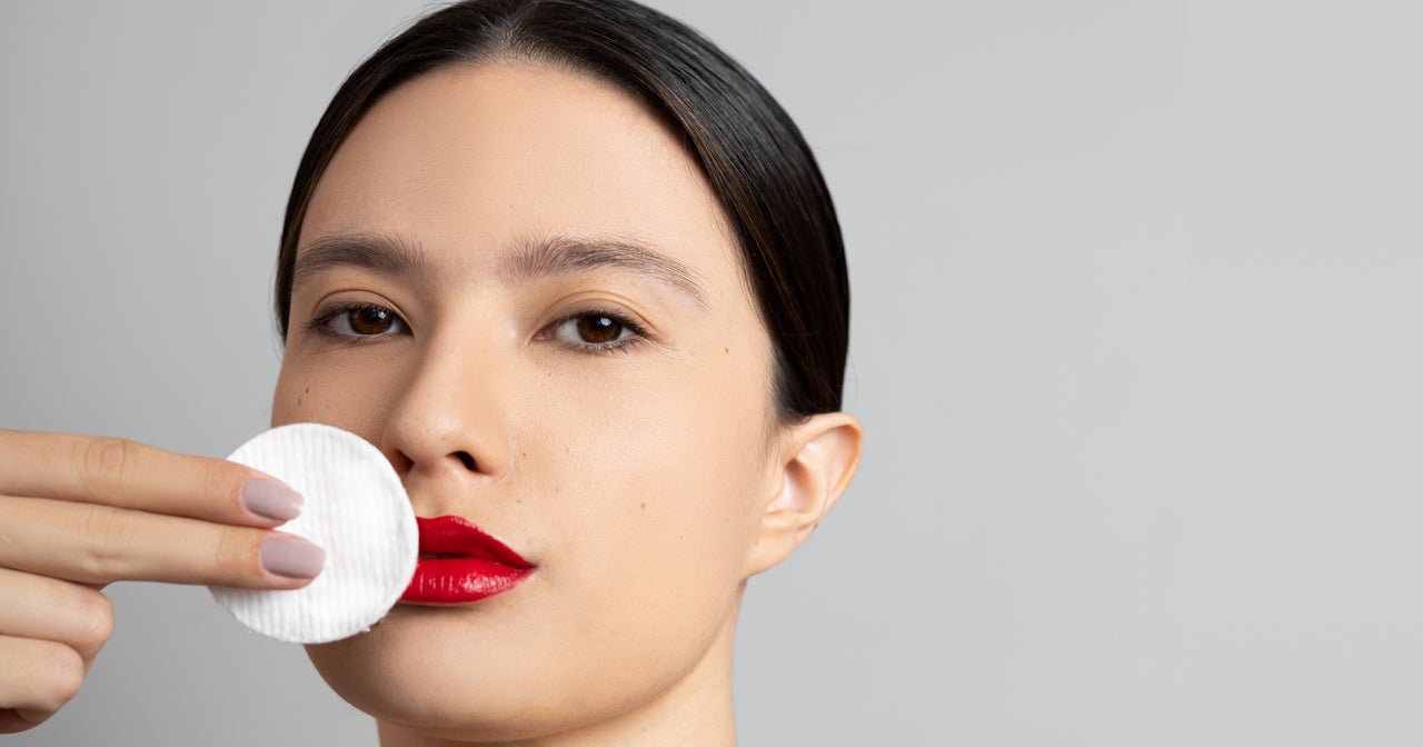 Close-up de uma mulher com lábios vermelhos vibrantes, usando um disco de algodão para remover a maquiagem. Ela está aplicando o demaquilante Dermotvin, que possui chá verde em sua formulação, para limpar suavemente a área da boca. A imagem destaca a importância da remoção da maquiagem na rotina de skincare e a eficácia de produtos com ingredientes naturais.