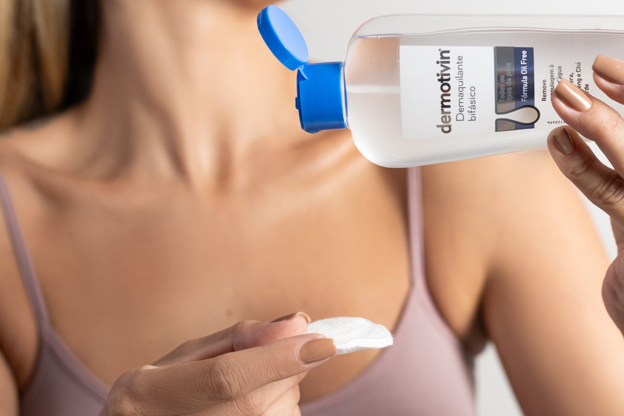Uma mulher está segurando um frasco de demaquilante bifásico Dermotivin com chá verde e um algodão. A mulher tem unhas pintadas de marrom claro.