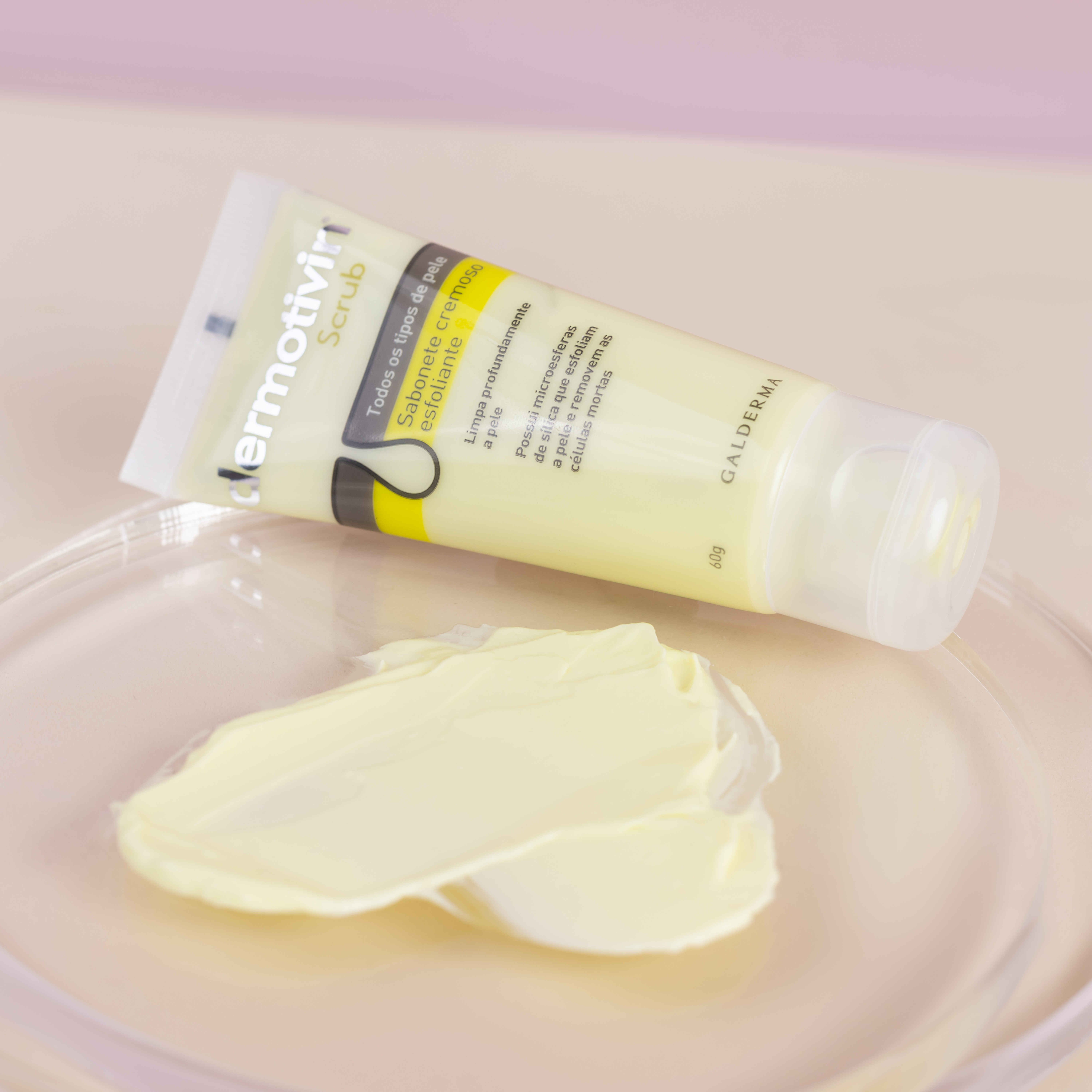 Close-up em ângulo inclinado do sabonete cremoso esfoliante Dermotivin Scrub. O tubo branco e amarelo está deitado sobre uma placa de vidro redonda. À frente do tubo, há uma amostra da textura do produto: um creme espesso de cor amarelo claro com pequenas partículas esfoliantes. Fundo rosa suave.