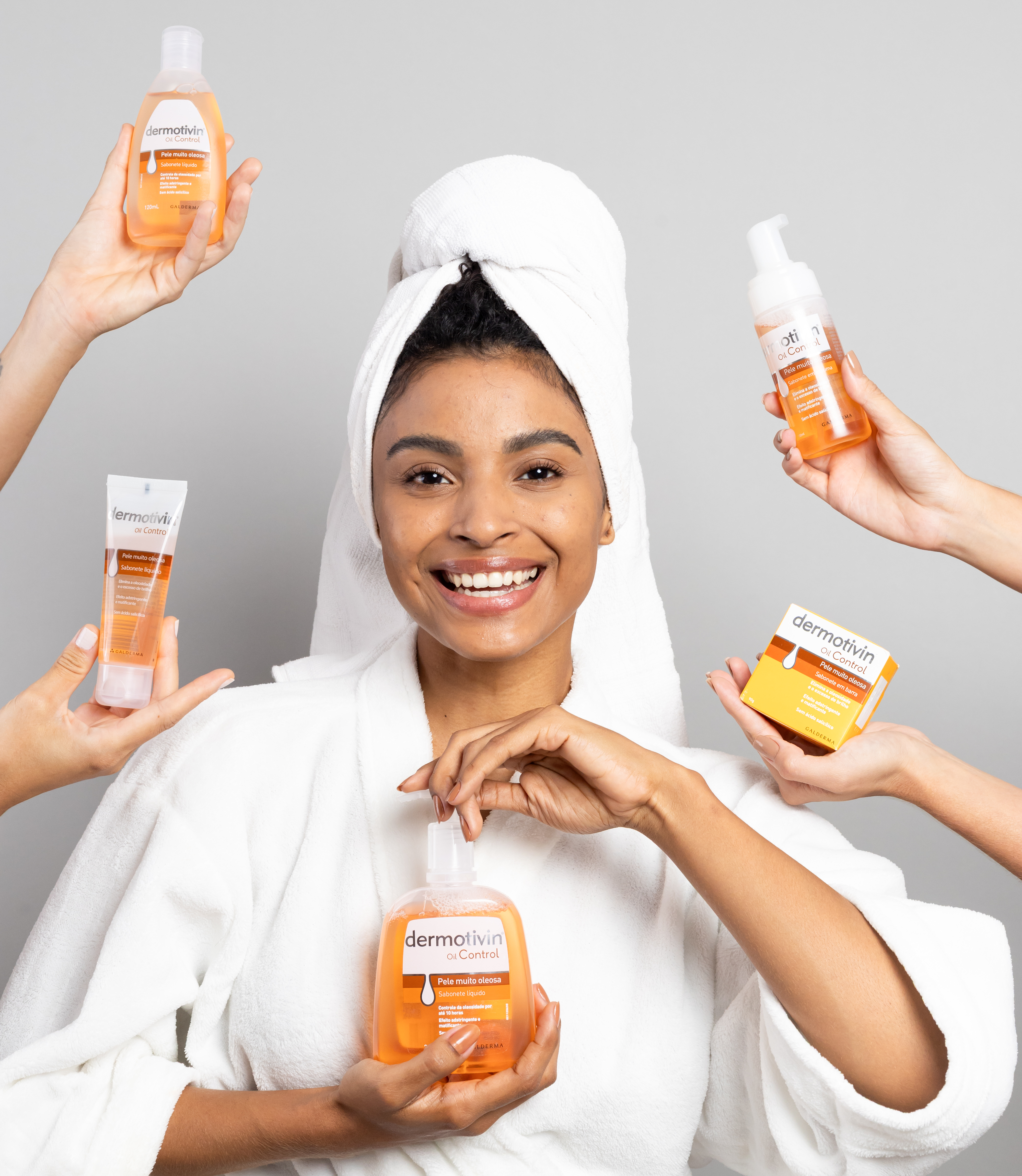 Uma mulher sorridente, com uma toalha na cabeça e roupão branco, está rodeada por várias mãos que oferecem produtos de limpeza facial da linha Dermotivin Oil Control. Os produtos, em tons de laranja e branco, incluem diferentes embalagens como frascos com pump, tubos e caixas, representando uma rotina completa de cuidados com a pele que utiliza a biotina para controle da oleosidade.