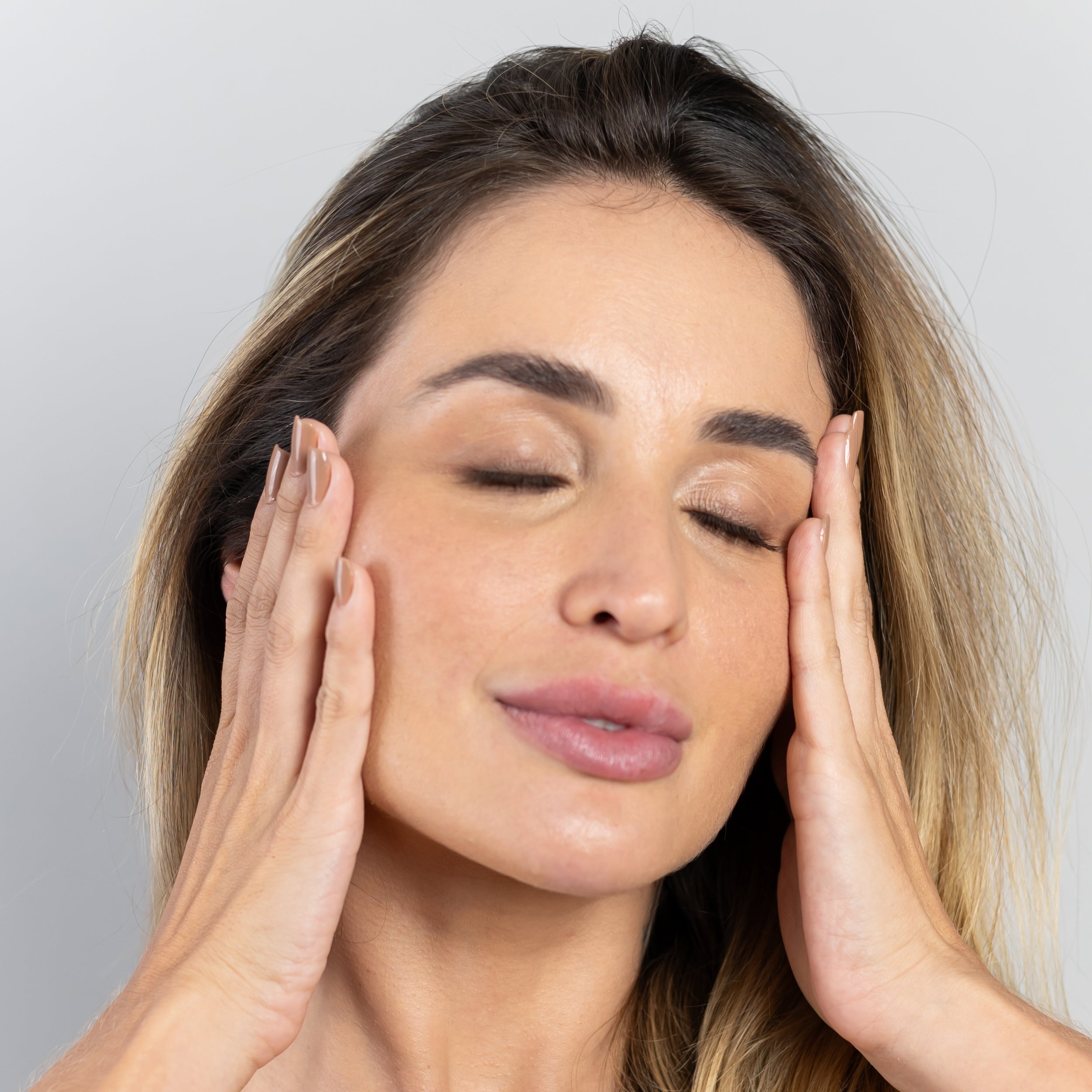 Close-up de uma mulher com os olhos fechados, as mãos gentilmente pressionadas nas laterais do rosto. Sua pele parece suave e bem cuidada, os cabelos loiros e castanhos emolduram seu rosto, e suas unhas estão bem tratadas, sugerindo os benefícios da biotina para a pele do rosto, cabelos e unhas.