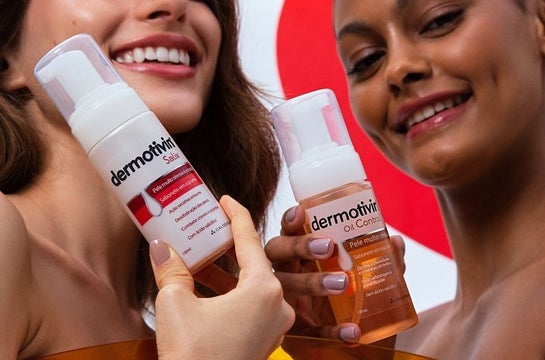 À esquerda, uma mulher branca sorri e segura embalagem de espuma de limpeza Dermotivin Salix. À direita, uma mulher negra sorri e segura embalagem da espuma de limpeza Dermotivin Oil Control.
