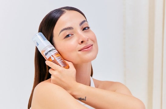 Mulher adulta branca usando maquiagem leve, para uma pele bonita, e segurando embalagem da espuma de limpeza Dermotivin Oil Control.