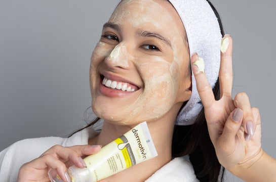 Uma mulher sorridente, vestindo um roupão branco e uma faixa de cabelo, aplica o Dermotivin Scrub no rosto. Ela segura o tubo do produto em uma mão e faz o sinal de "paz e amor" com a outra, transmitindo uma sensação de bem-estar e cuidado com a pele. A imagem destaca a aplicação do produto de esfoliação facial, mostrando a textura cremosa e a cobertura uniforme na pele. A expressão feliz da modelo reforça a ideia de que a esfoliação facial com Dermotivin Scrub é um momento agradável e parte de uma rotina d