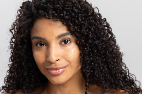 Mulher jovem com cabelos cacheados escuros e pele radiante sorrindo suavemente, demonstrando uma pele saudável e bem cuidada em fundo neutro.