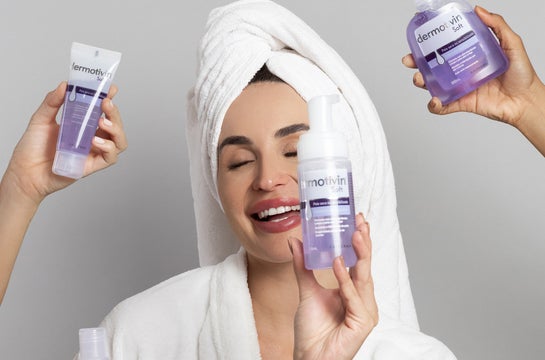 Uma mulher com uma toalha enrolada na cabeça está sorrindo e segurando um frasco de produto para cuidados com a pele. Ela está vestindo um roupão branco. Três mãos seguram outros produtos para cuidados com a pele ao redor dela. Os produtos estão todos em embalagens roxas e brancas. O fundo é cinza liso. A imagem geral tem uma estética limpa e brilhante, sugerindo um foco em cuidados com a pele e beleza.