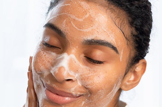 Mulher de pele negra com os olhos fechados aplicando sabonete facial espumante na zona T do rosto (testa, nariz e queixo). A espuma branca cobre principalmente a testa e o nariz, destacando a área onde a oleosidade costuma ser mais intensa.