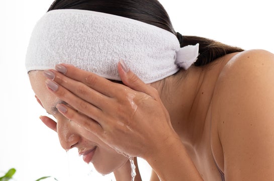 Mulher realizando sua rotina de cuidados faciais, lavando o rosto com água sobre uma pia. Ela usa uma faixa branca para prender o cabelo, mantendo-o afastado do rosto durante a limpeza. A imagem mostra o momento da enxágue, com a água escorrendo suavemente pelas mãos posicionadas sobre o rosto, demonstrando a importância da limpeza adequada na rotina de skincare para pele oleosa.