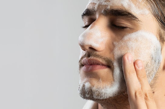 Um close-up de um homem jovem com cabelo escuro e barba por fazer, de perfil, com os olhos fechados, aplicando Espuma de Limpeza no rosto. Ele está usando uma faixa de cabelo preta e uma toalha branca. A espuma branca cobre a testa, bochecha e queixo, enquanto ele usa o dedo indicador e médio da mão direita para espalhar o produto na bochecha. O fundo é um cinza claro e desfocado.