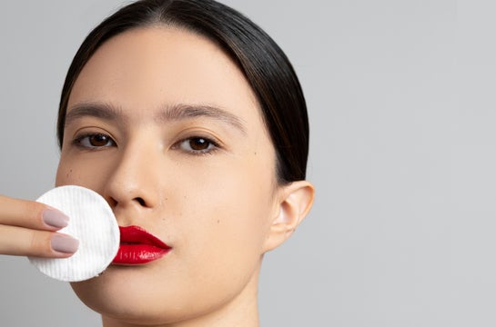 Close-up de uma mulher com lábios vermelhos vibrantes, usando um disco de algodão para remover a maquiagem. Ela está aplicando o demaquilante Dermotvin, que possui chá verde em sua formulação, para limpar suavemente a área da boca. A imagem destaca a importância da remoção da maquiagem na rotina de skincare e a eficácia de produtos com ingredientes naturais.