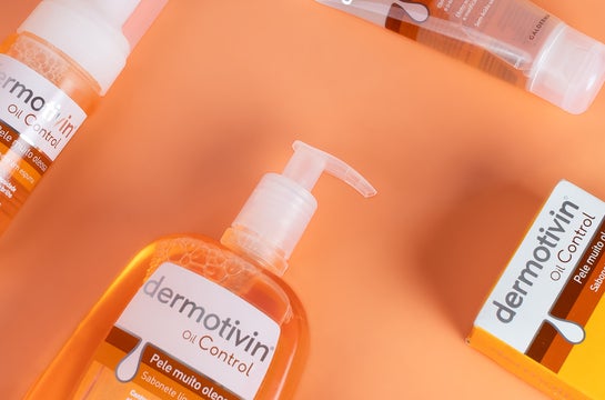Linha completa Dermotivin Oil Control para pele oleosa disposta sobre fundo laranja vibrante com detalhes amarelos. Produtos em diferentes formatos: sabonete líquido em frasco pump e sabonete em espuma, todos com embalagem branca e detalhes laranjas.