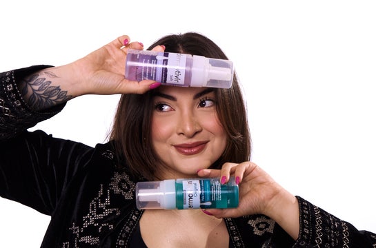 Influenciadora sorrindo segura dois frascos de Espuma de Limpeza Facial Dermotivin, um roxo, da linha Soft, e um azul, da linha Original, posicionando um sobre a testa e outro próximo ao queixo.