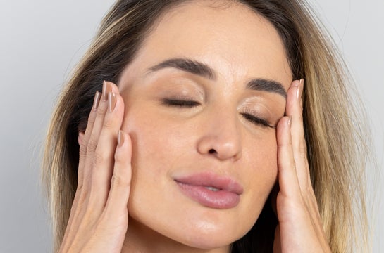 Close-up de uma mulher com os olhos fechados, as mãos gentilmente pressionadas nas laterais do rosto. Sua pele parece suave e bem cuidada, os cabelos loiros e castanhos emolduram seu rosto, e suas unhas estão bem tratadas, sugerindo os benefícios da biotina para a pele do rosto, cabelos e unhas.